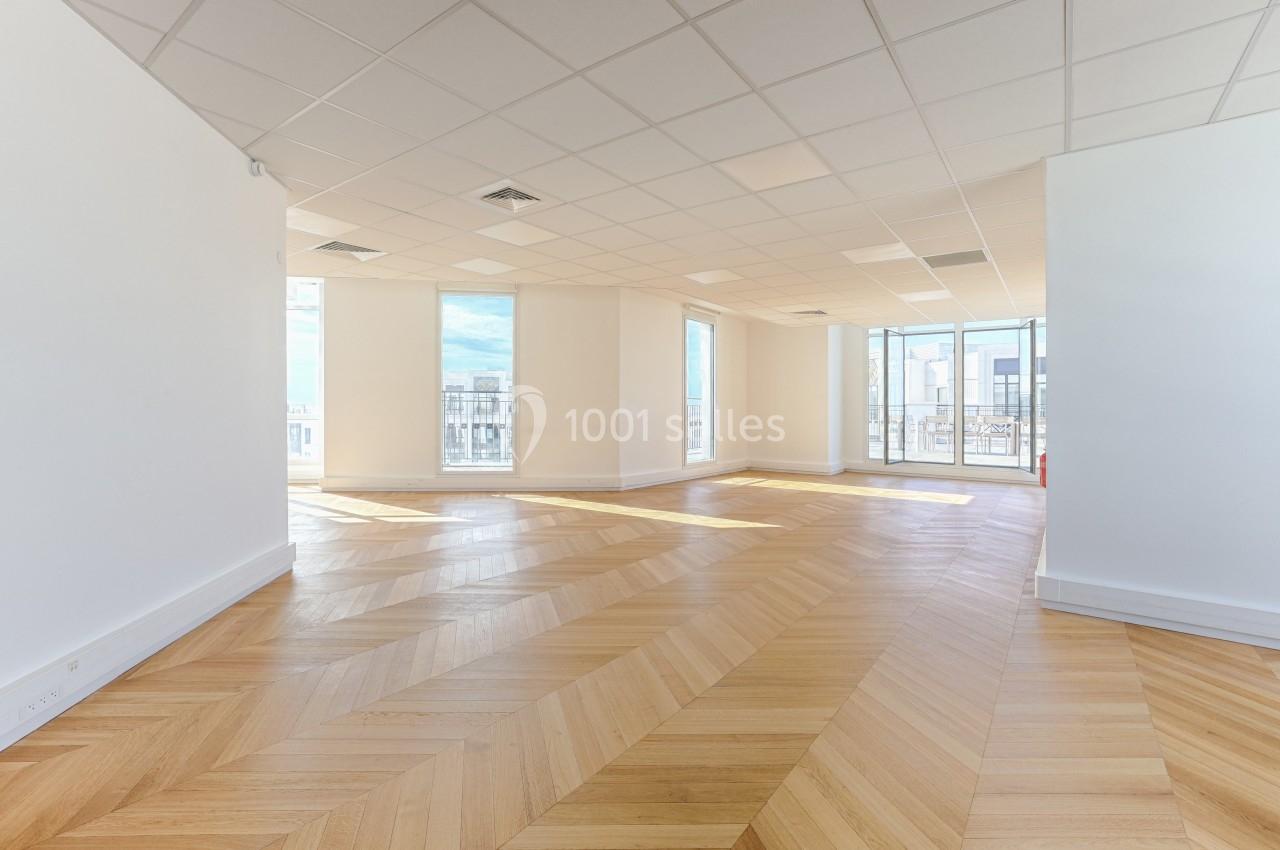Bureau lumineux vide avec parquet en chevrons, grandes fenêtres et plafond avec dalles blanches.