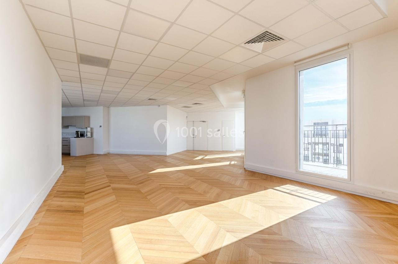 Salle lumineuse avec parquet en chevrons, plafond à dalles blanches, cuisine ouverte et grande fenêtre donnant sur un balcon.