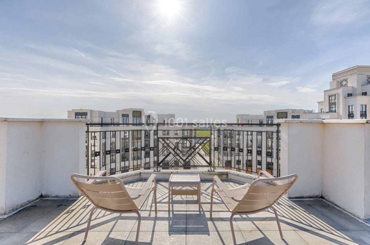 Deux chaises et une table sur une terrasse ensoleillée avec vue sur des bâtiments modernes et un ciel dégagé.