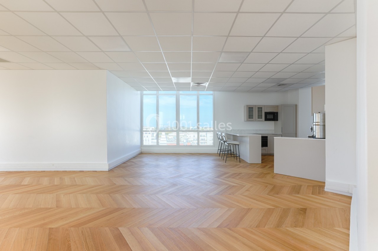 Pièce lumineuse avec parquet en chevrons, grande fenêtre, cuisine ouverte et plafond à dalles blanches.