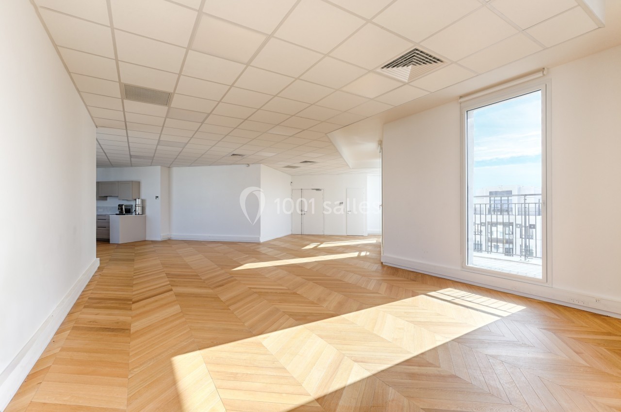 Pièce lumineuse avec parquet en bois clair, plafond à dalles blanches, cuisine ouverte et grande fenêtre donnant sur un…