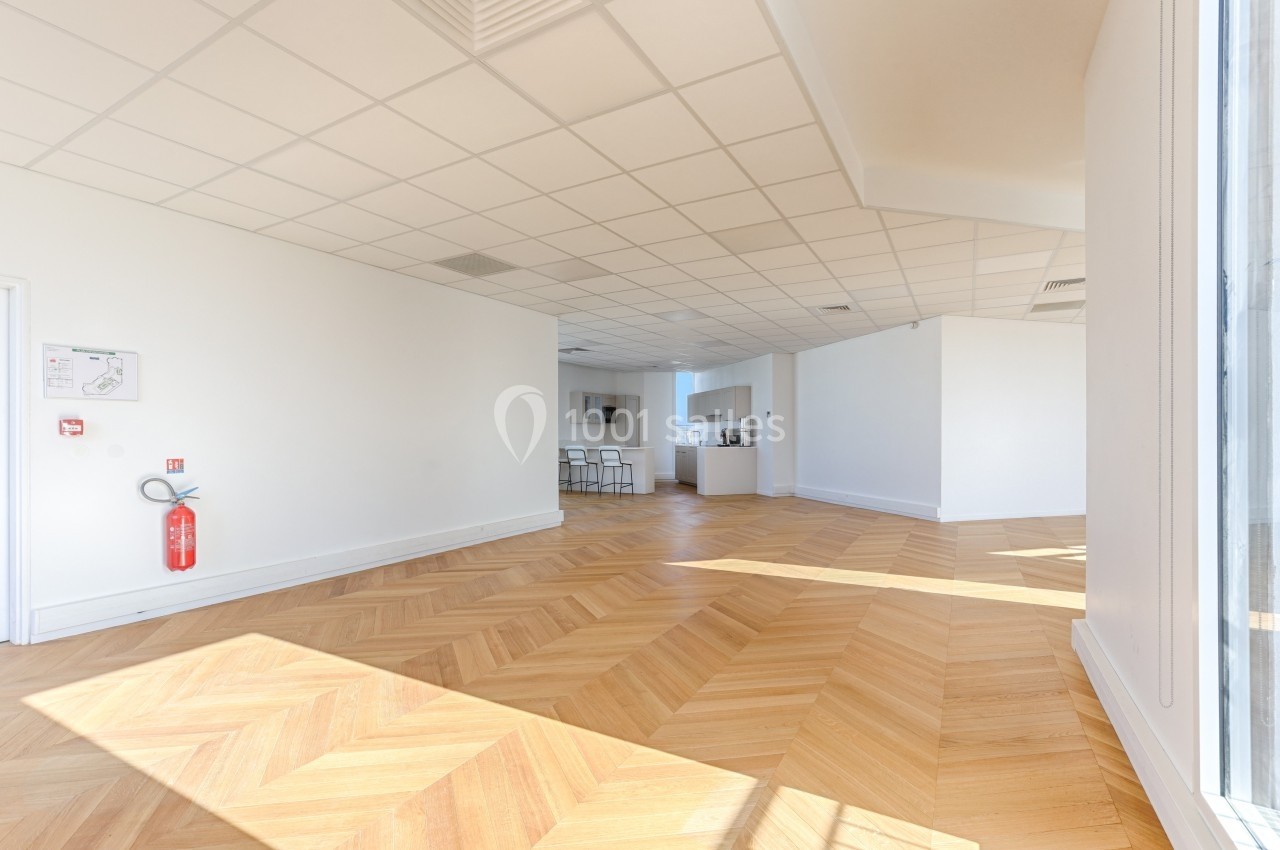 Salle lumineuse avec parquet en chevrons, murs blancs, cuisine ouverte et mobilier minimaliste en arrière-plan.