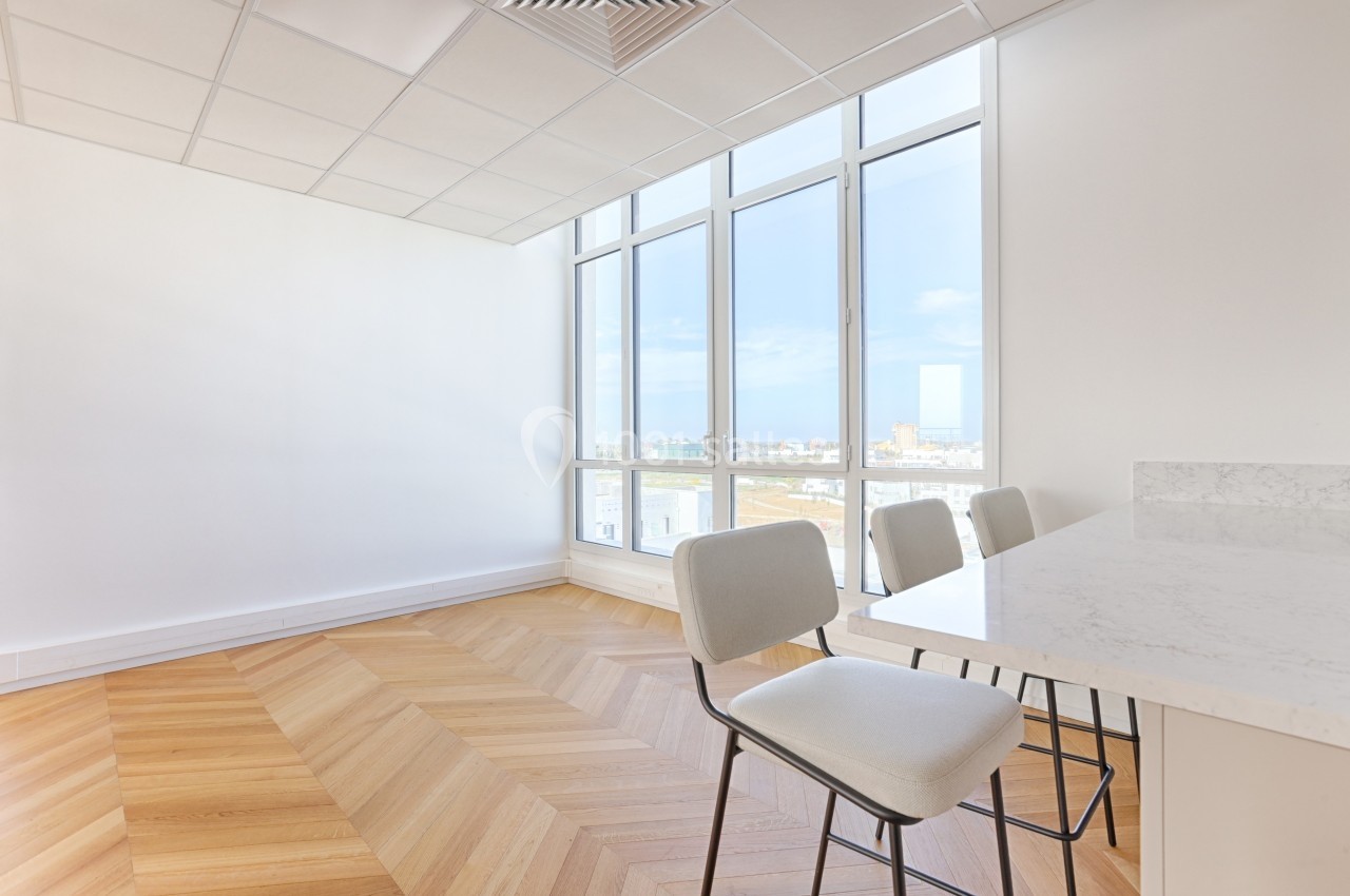 Salle lumineuse avec parquet en bois, grandes fenêtres donnant sur une vue dégagée, et comptoir avec chaises.