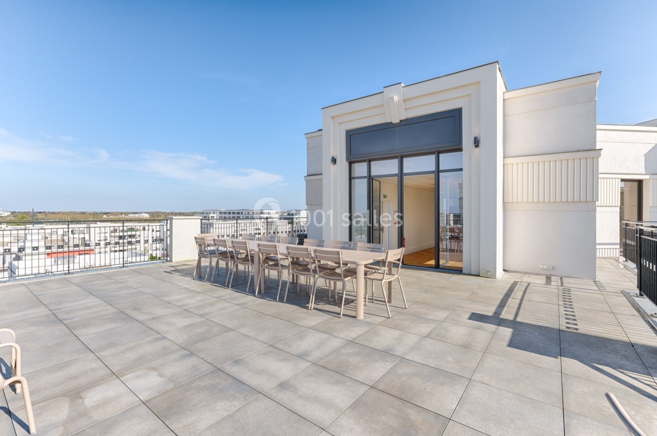 Grande terrasse ensoleillée avec table et chaises, donnant sur un bâtiment moderne et un paysage urbain lointain.
