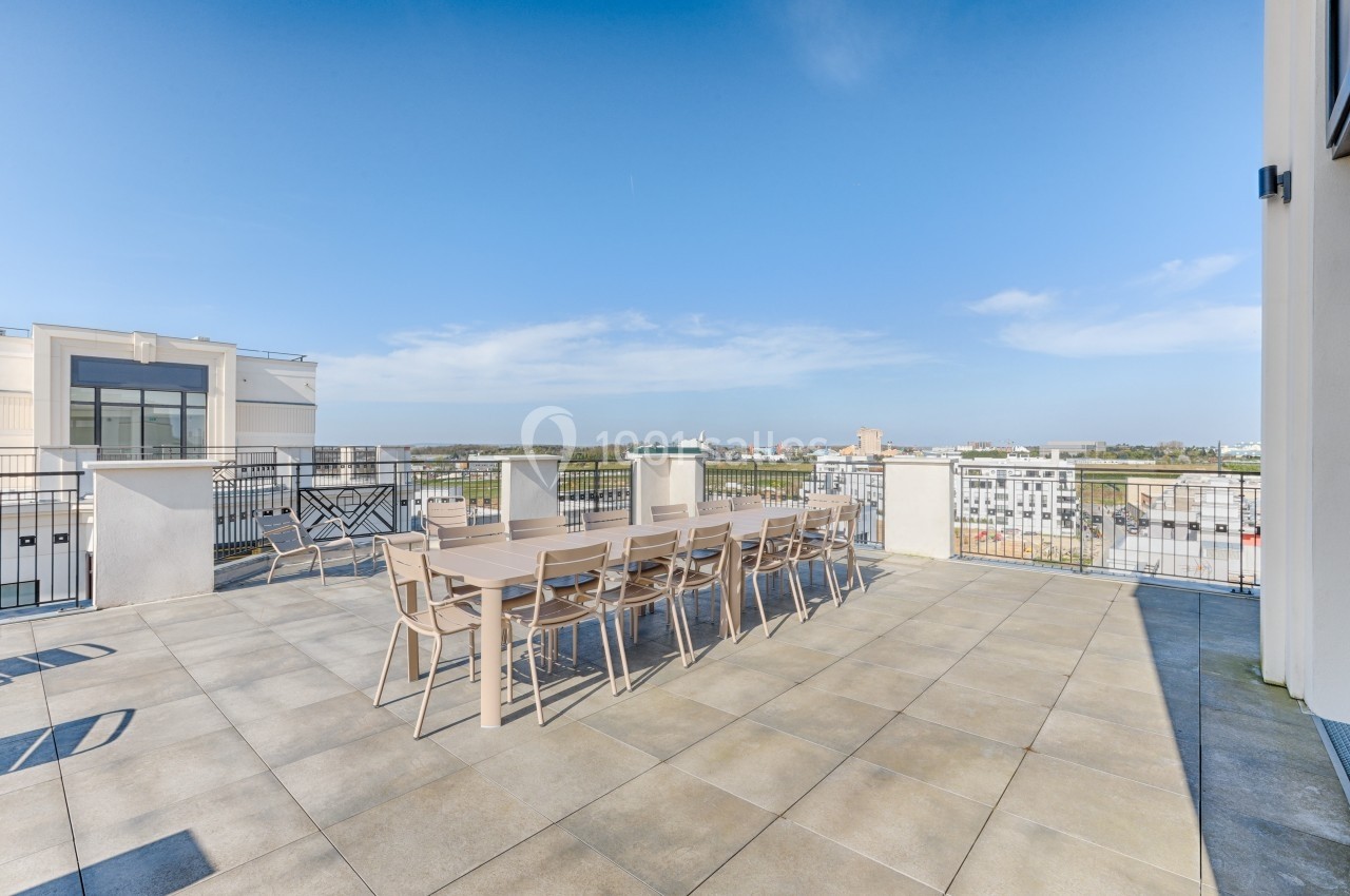 Terrasse spacieuse avec grande table en bois et chaises, offrant une vue dégagée sur des bâtiments et le ciel.