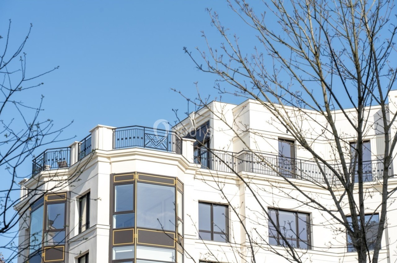 Façade d'un immeuble moderne avec balcons et grandes fenêtres, partiellement masquée par des branches d'arbres.