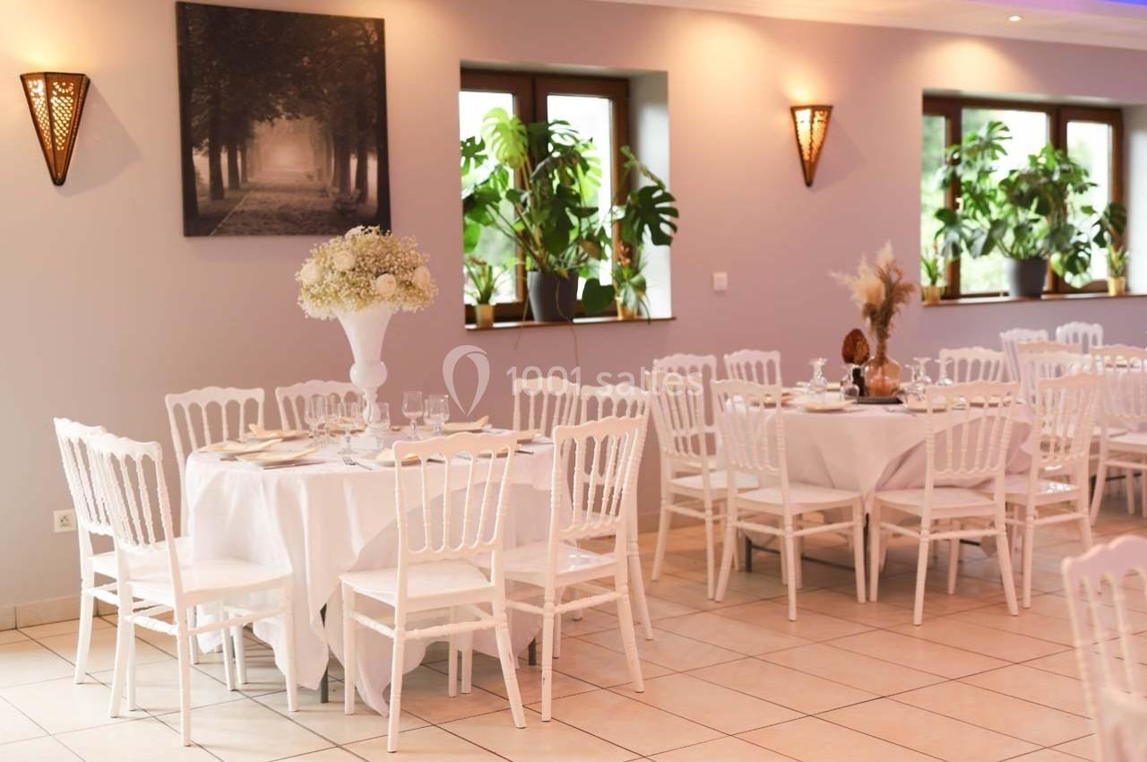 Salle de réception lumineuse avec tables rondes dressées, chaises blanches et décorations florales.