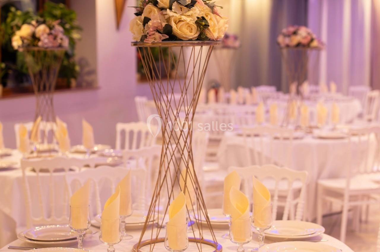 Centre de table floral sur un support doré, entouré de tables dressées avec nappes blanches et serviettes pliées.