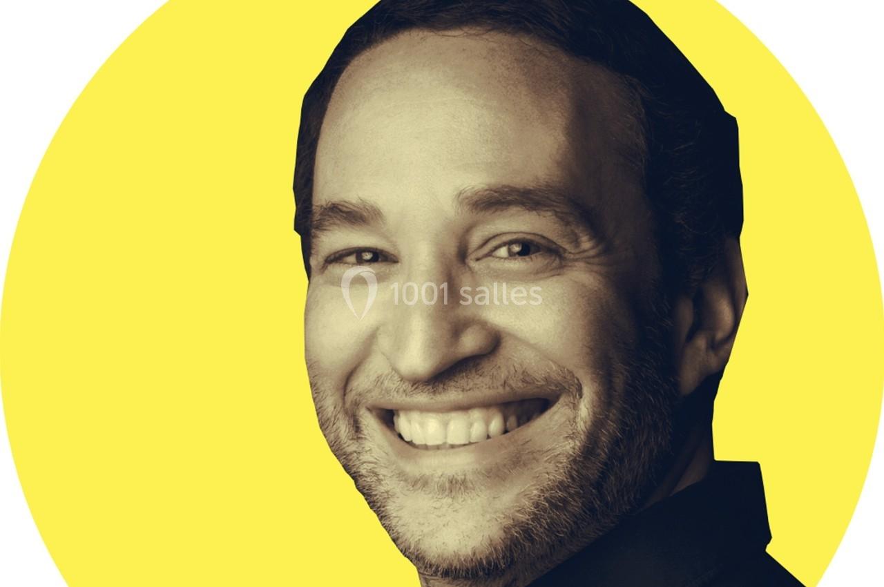 Portrait d'un homme souriant avec une barbe légère, sur un fond jaune uni.