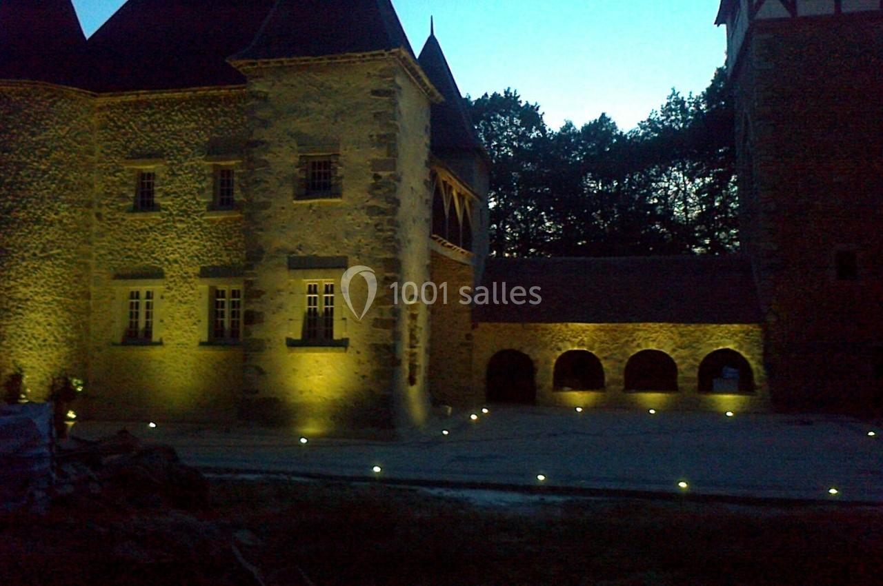 Façade d'un château en pierre éclairée par des lumières au sol, avec une cour et des arbres en arrière-plan.