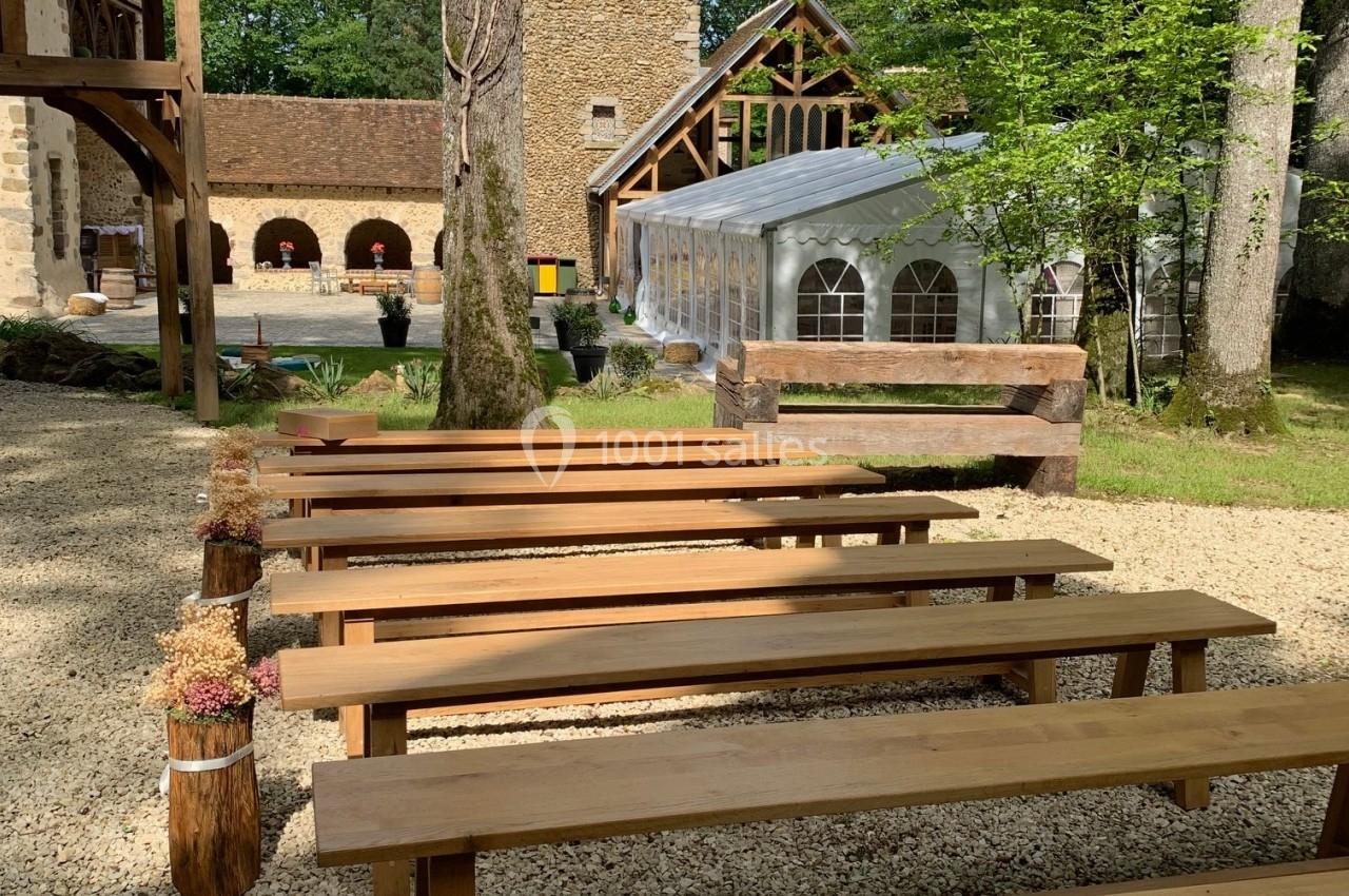 Bancs en bois alignés en extérieur, devant une tour en pierre et une structure avec toit transparent, entourés d'arbres.