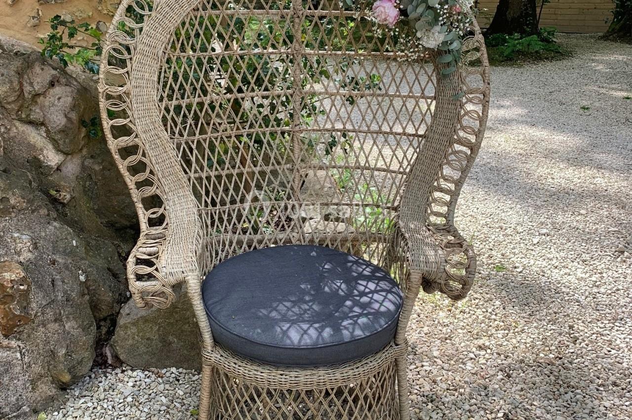 Fauteuil en rotin avec coussin noir, décoré de fleurs, placé sur un sol en gravier dans un jardin arboré.