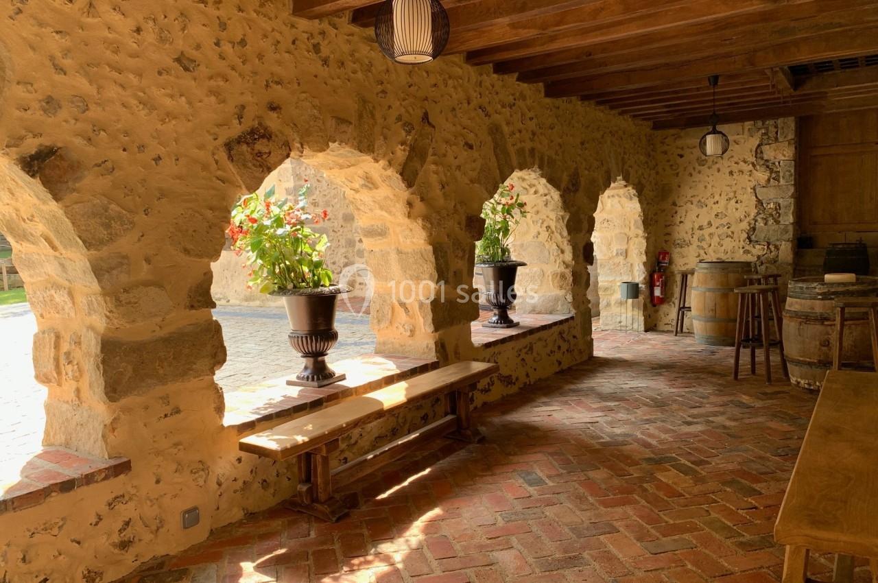 Arcades en pierre avec pots de fleurs, éclairées par la lumière naturelle, sur un sol en briques dans un espace rustique.