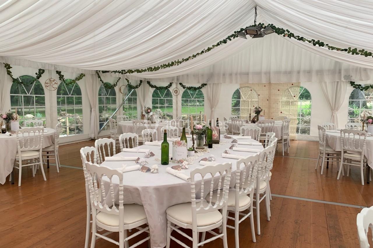 Intérieur d'une tente blanche décorée pour un événement, avec tables rondes dressées et chaises blanches sur un sol en bois.
