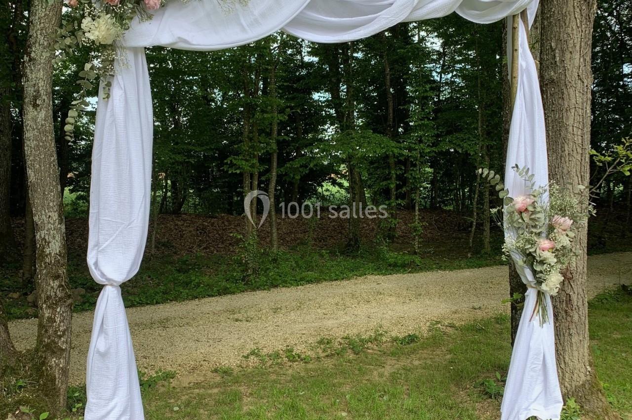 Arche de mariage en bois décorée de drapés blancs et de fleurs roses et blanches, installée dans un cadre boisé.
