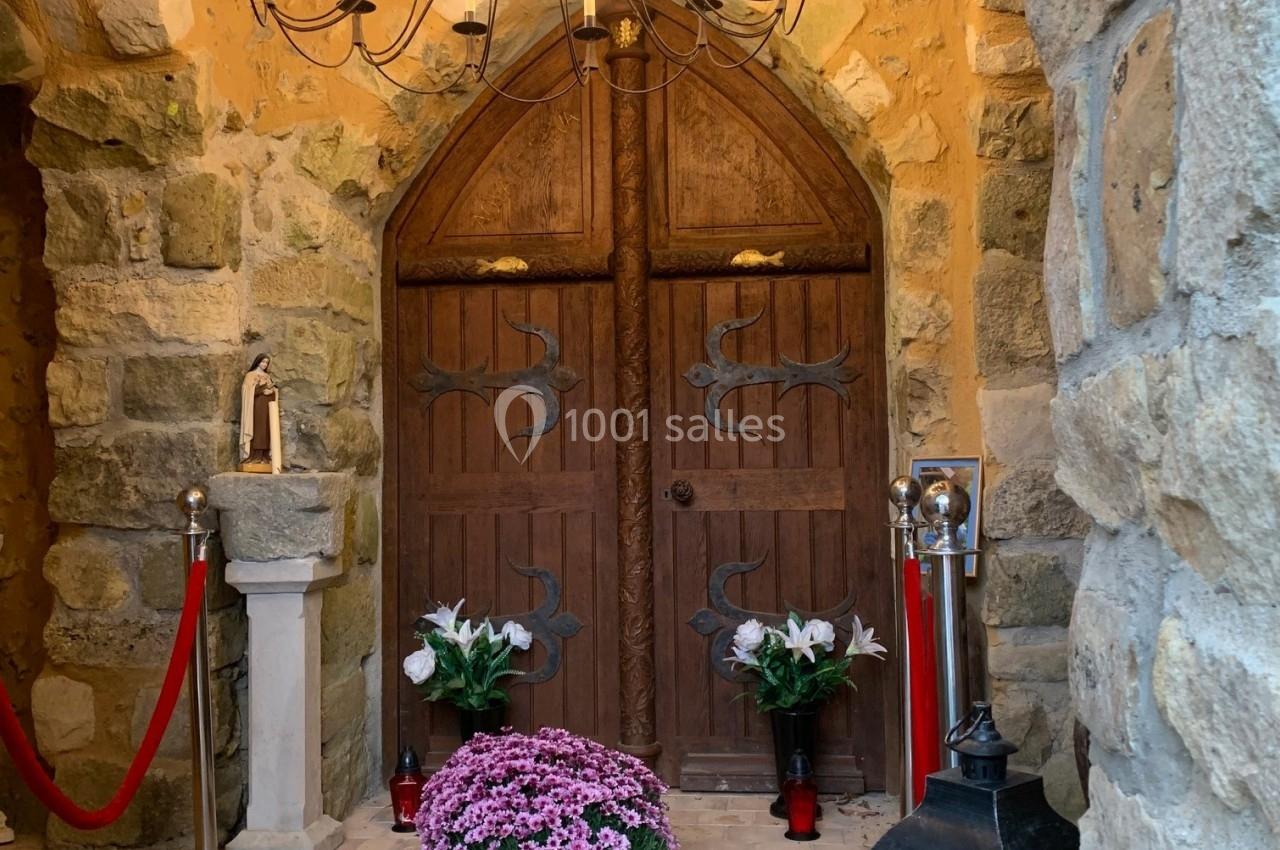 Porte en bois sculptée entourée de murs en pierre, décorée de fleurs, de chandeliers et d'une statue religieuse.