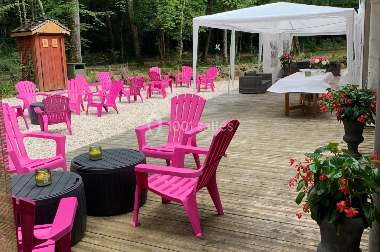 Terrasse en bois avec chaises roses, tables noires, tonnelle blanche et végétation dense en arrière-plan.