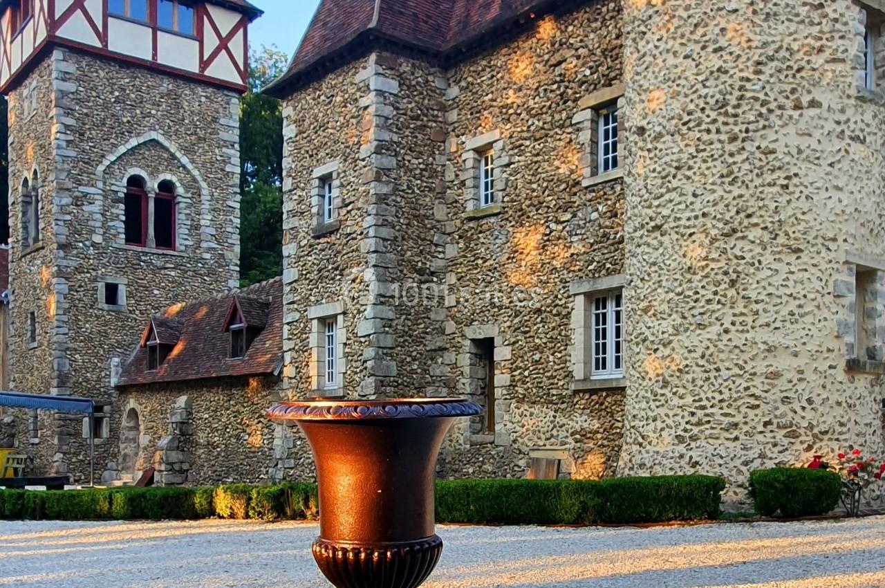 Château en pierre avec tours et toits en ardoise, éclairé par une lumière douce, avec une urne décorative au premier plan.