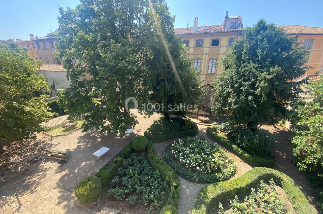 Jardin aménagé avec des parterres de buis, des bancs et des arbres, entouré de bâtiments anciens sous un ciel dégagé.