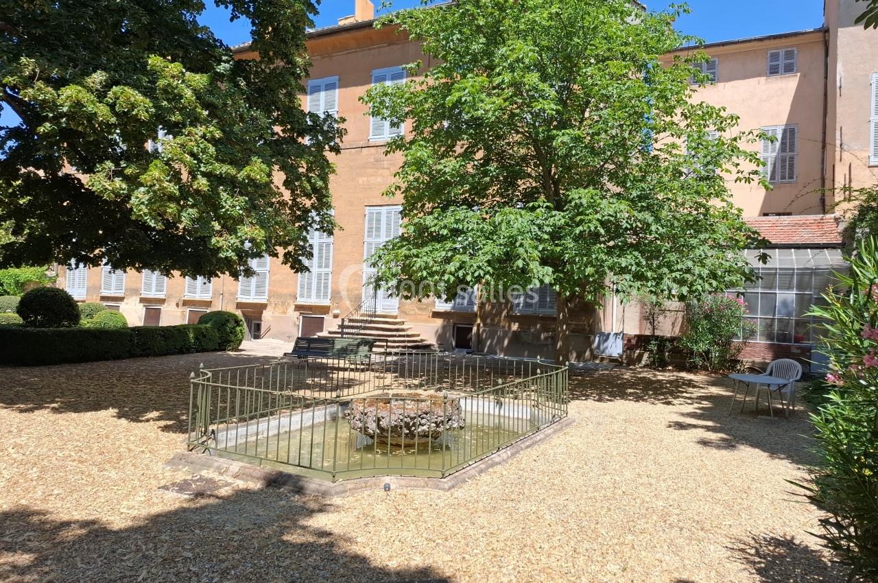 Cour avec un petit bassin entouré de graviers, bordée d'arbres et d'un bâtiment ancien aux volets blancs.