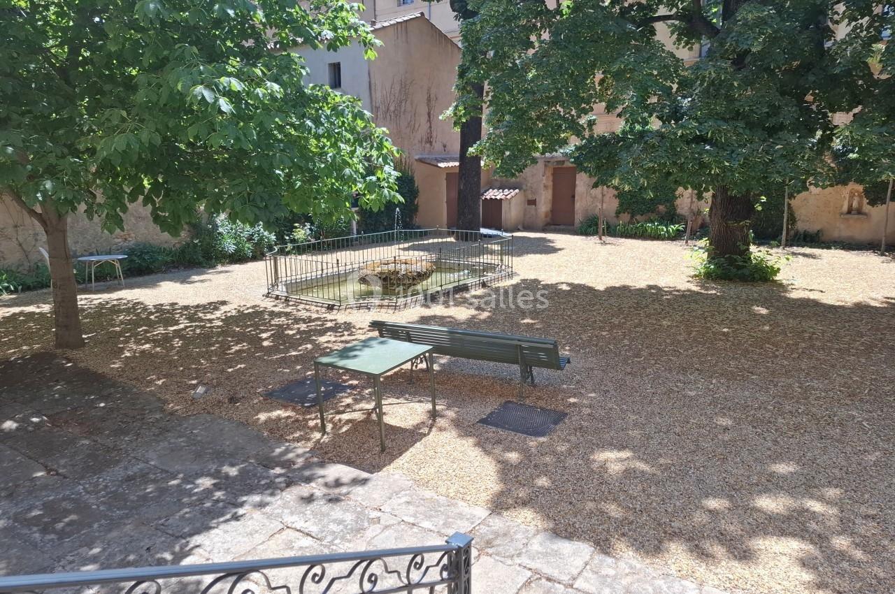 Petit jardin ombragé avec bancs, table en métal, arbres et un petit bassin central entouré d'une grille.