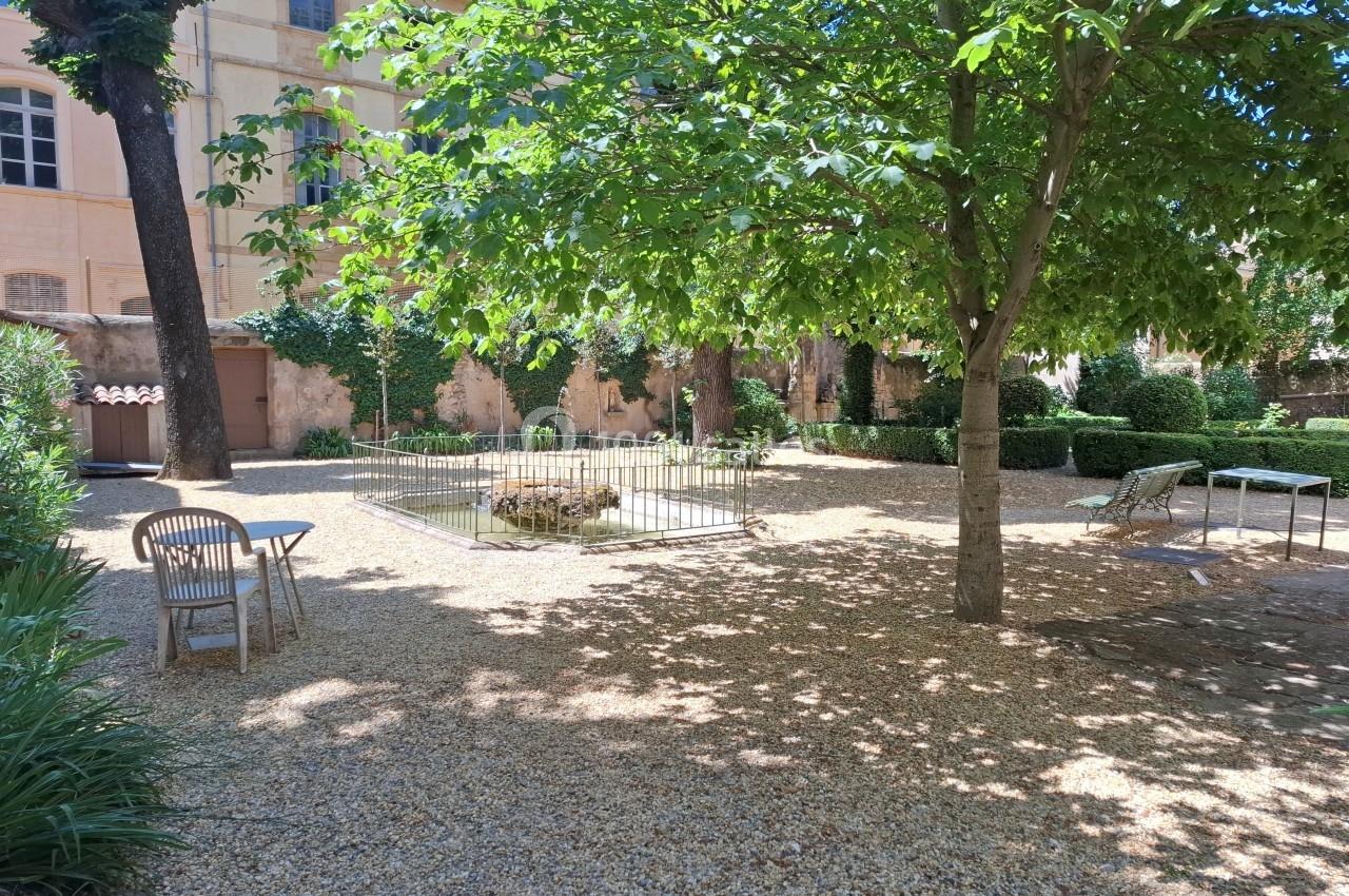 Jardin ombragé avec des arbres, des chaises en bois, une table et une petite clôture entourant un espace central.