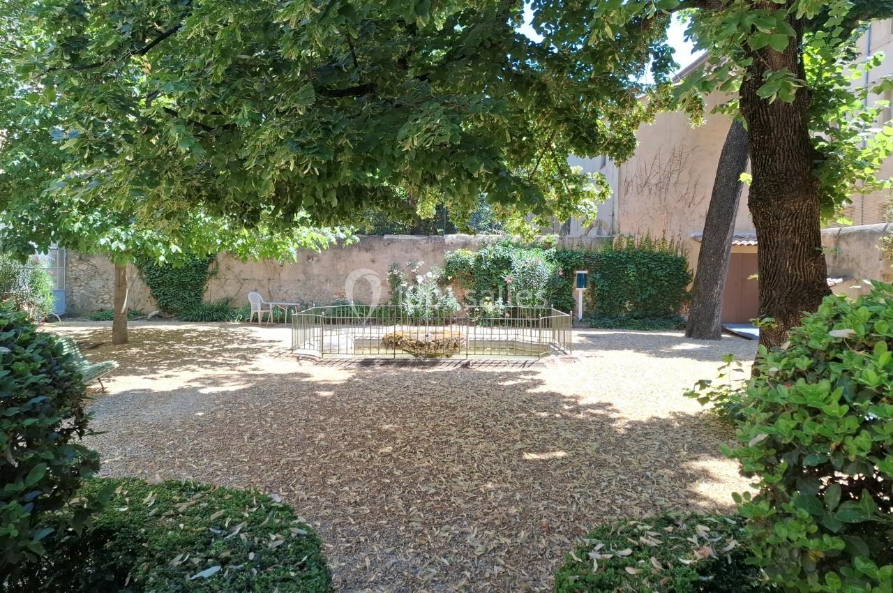 Cour ombragée avec des arbres, un parterre central entouré de barrières et des murs en pierre en arrière-plan.