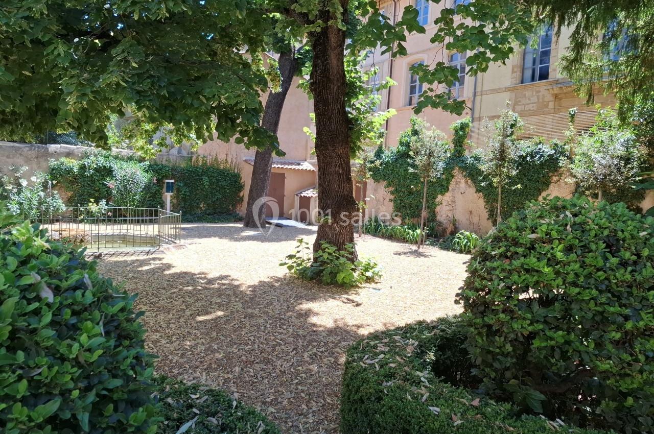 Cour ombragée avec arbres, buissons taillés et murs en pierre, entourée de végétation et baignée de lumière naturelle.