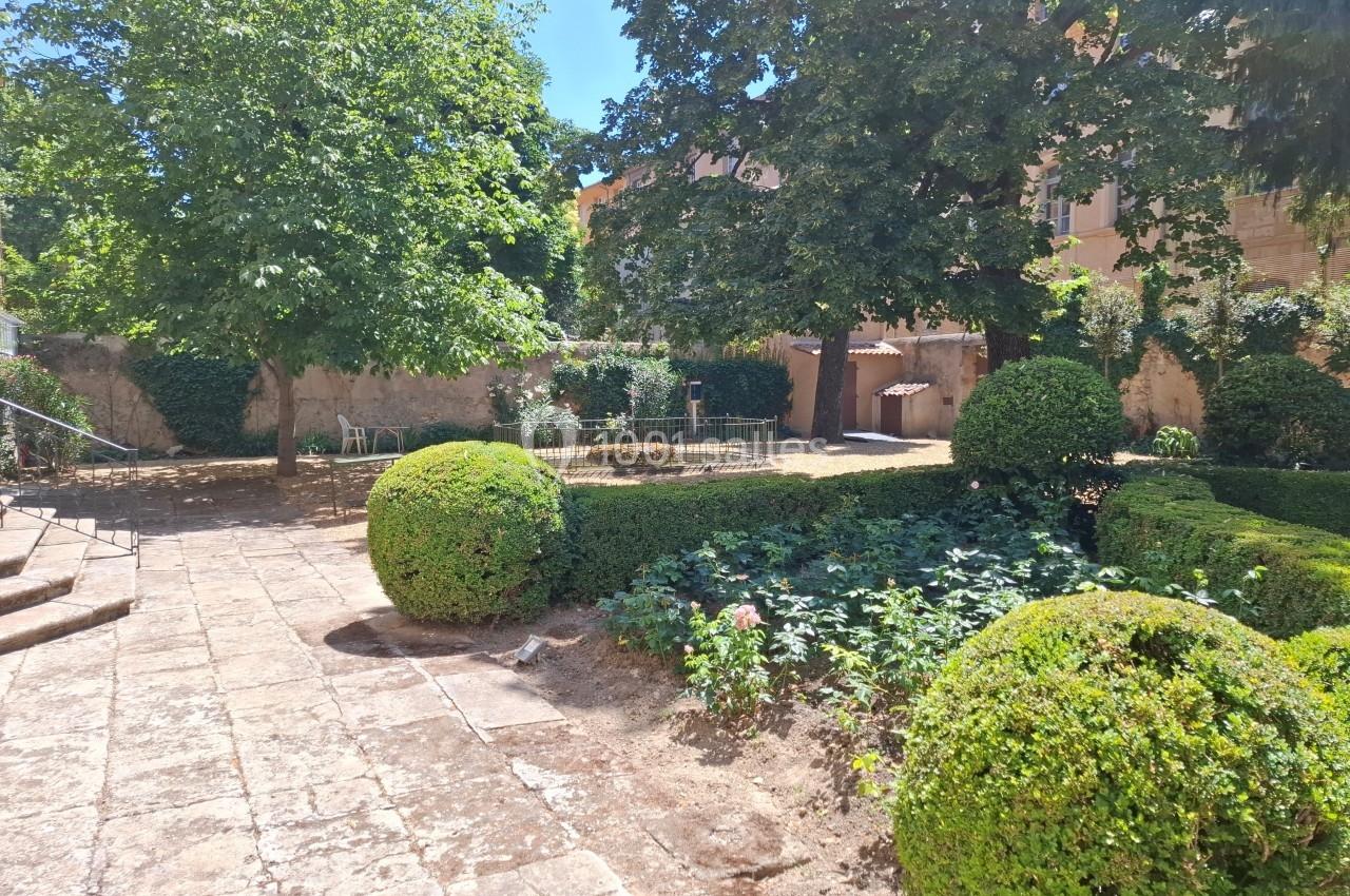 Jardin ombragé avec arbres, buissons taillés, parterre fleuri et mobilier de jardin près d'un bâtiment en arrière-plan.