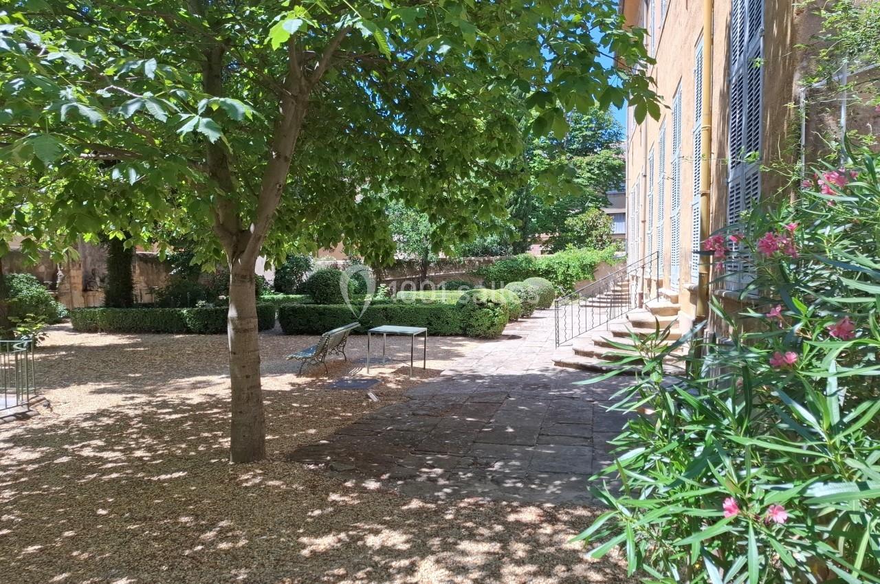 Cour ombragée avec arbres, buissons taillés, table et chaises en métal, bordée par un bâtiment aux volets bleus.