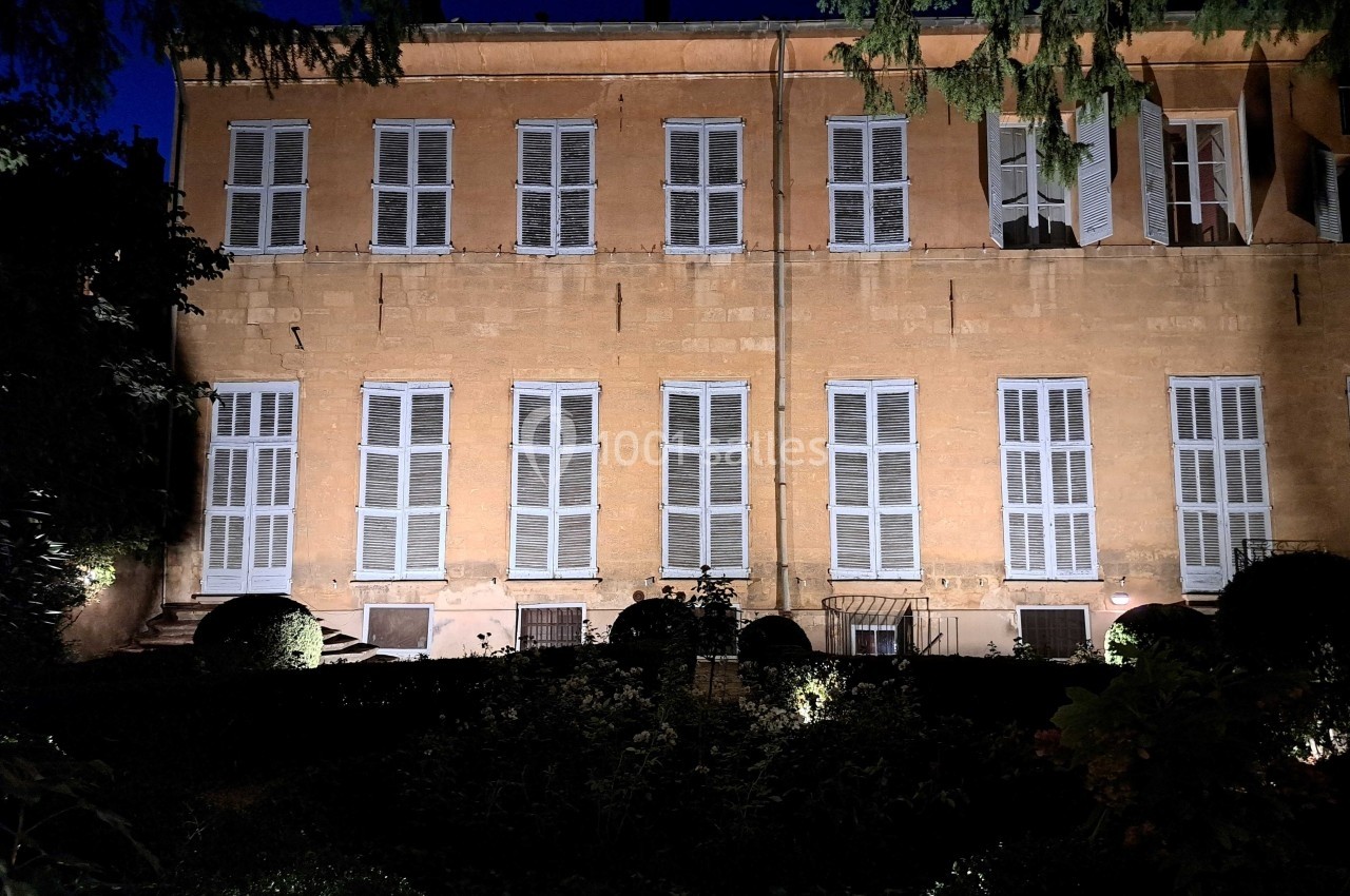 Façade d'un bâtiment ancien en pierre avec volets fermés, éclairée la nuit, entourée de végétation.