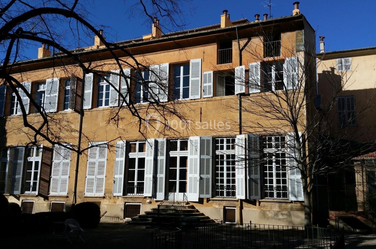 Façade d'un bâtiment ancien avec volets blancs, grandes fenêtres et arbres sans feuilles au premier plan.