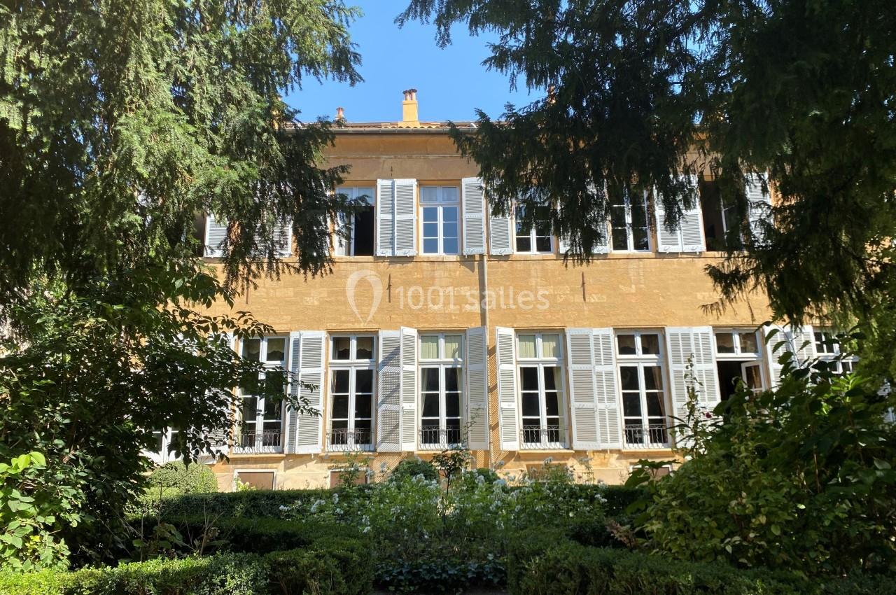 Façade d'un bâtiment ancien en pierre dorée avec volets blancs, entouré de végétation et d'arbustes.