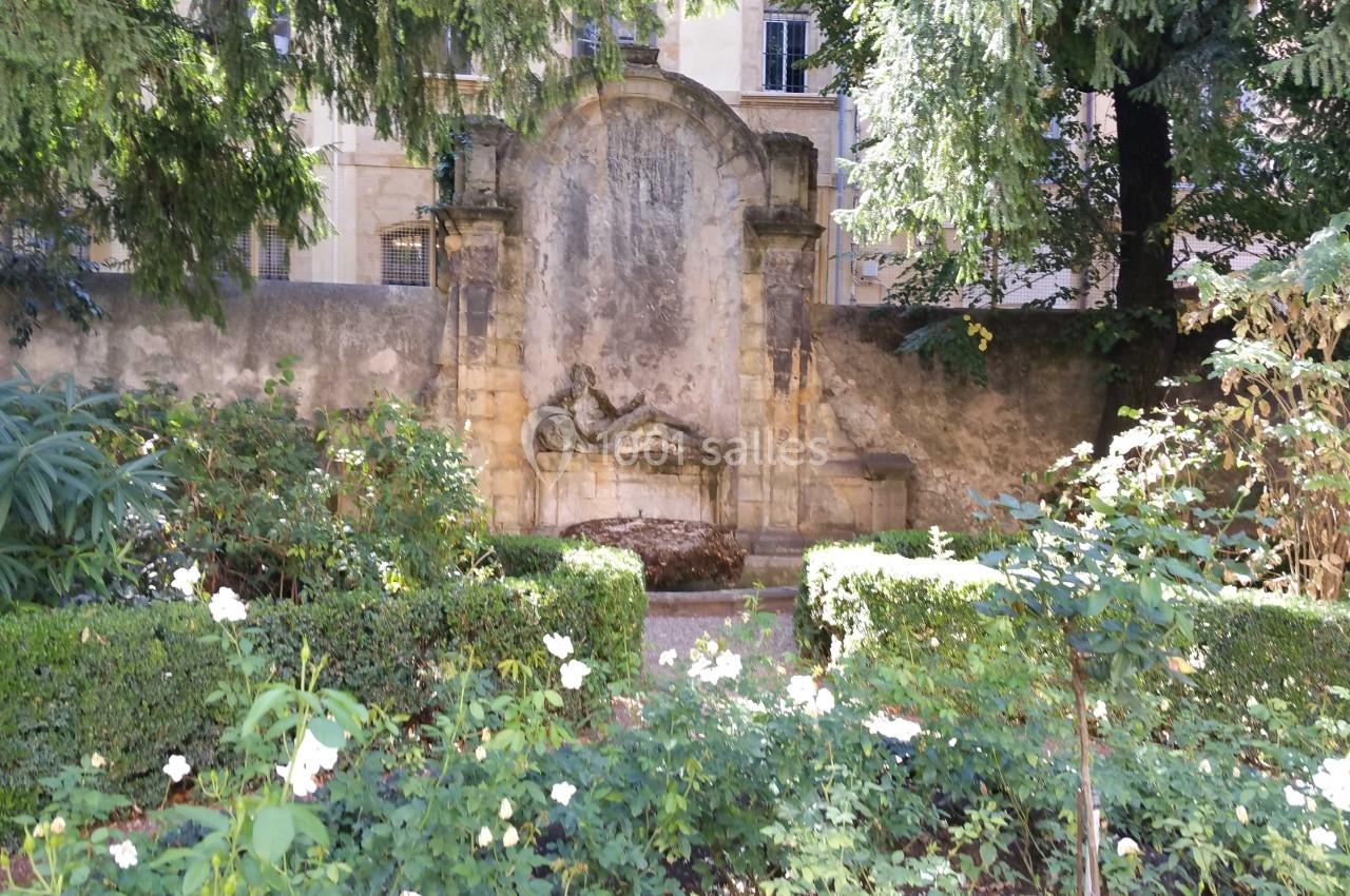Fontaine en pierre ancienne entourée de verdure et de fleurs blanches dans un jardin ombragé.