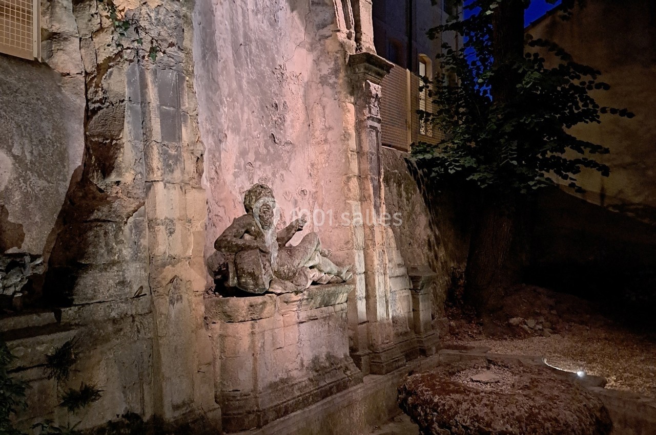 Fontaine en pierre ancienne éclairée la nuit, entourée d'arbres et de végétation dans un cadre urbain.