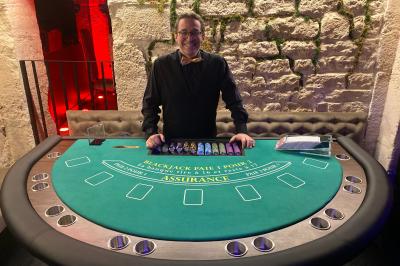 Table de jeu de casino avec tapis vert numéroté et porte-gobelets intégrés, entourée de personnes debout.