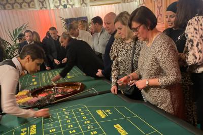 Table de jeu de casino avec tapis vert numéroté et porte-gobelets intégrés, entourée de personnes debout.