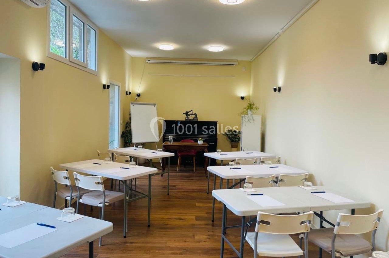 Salle lumineuse avec tables et chaises disposées pour une réunion ou une formation, tableau blanc et plantes en arrière-plan.