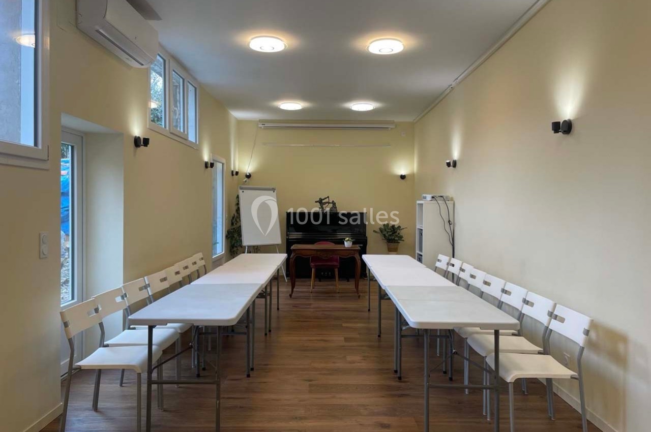 Salle lumineuse avec tables blanches disposées en U, chaises alignées, tableau blanc et piano en arrière-plan.