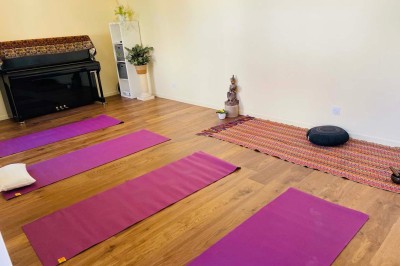 Salle lumineuse avec des tapis de yoga violets alignés sur un sol en bois, un piano et des décorations en arrière-plan.