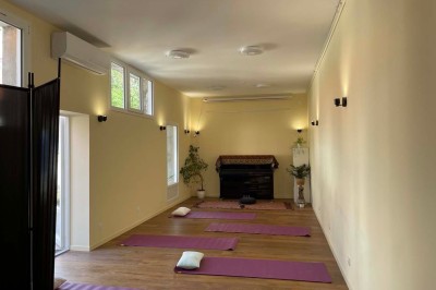 Salle lumineuse avec des tapis de yoga violets alignés sur un sol en bois, un piano et des décorations en arrière-plan.