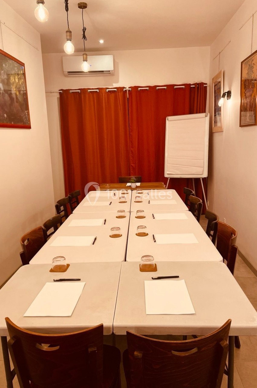 Salle de réunion avec tables alignées, chaises, blocs-notes, stylos, rideaux rouges et un tableau de conférence.