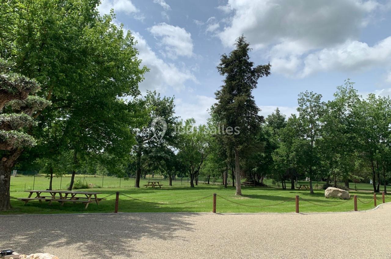 Espace vert avec pelouse, arbres, tables de pique-nique et ciel partiellement nuageux en arrière-plan.