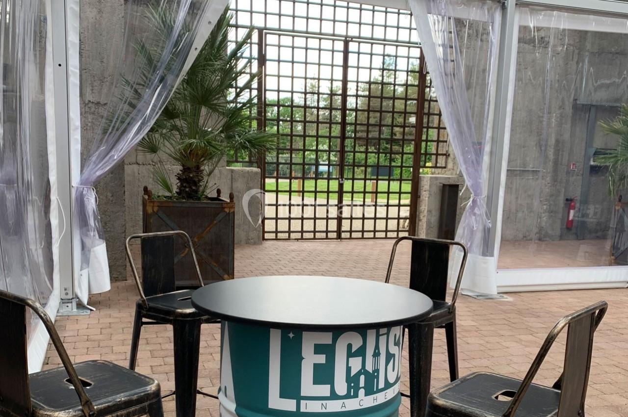 Table ronde et chaises en métal disposées sous une structure transparente, avec des plantes et une grille en arrière-plan.