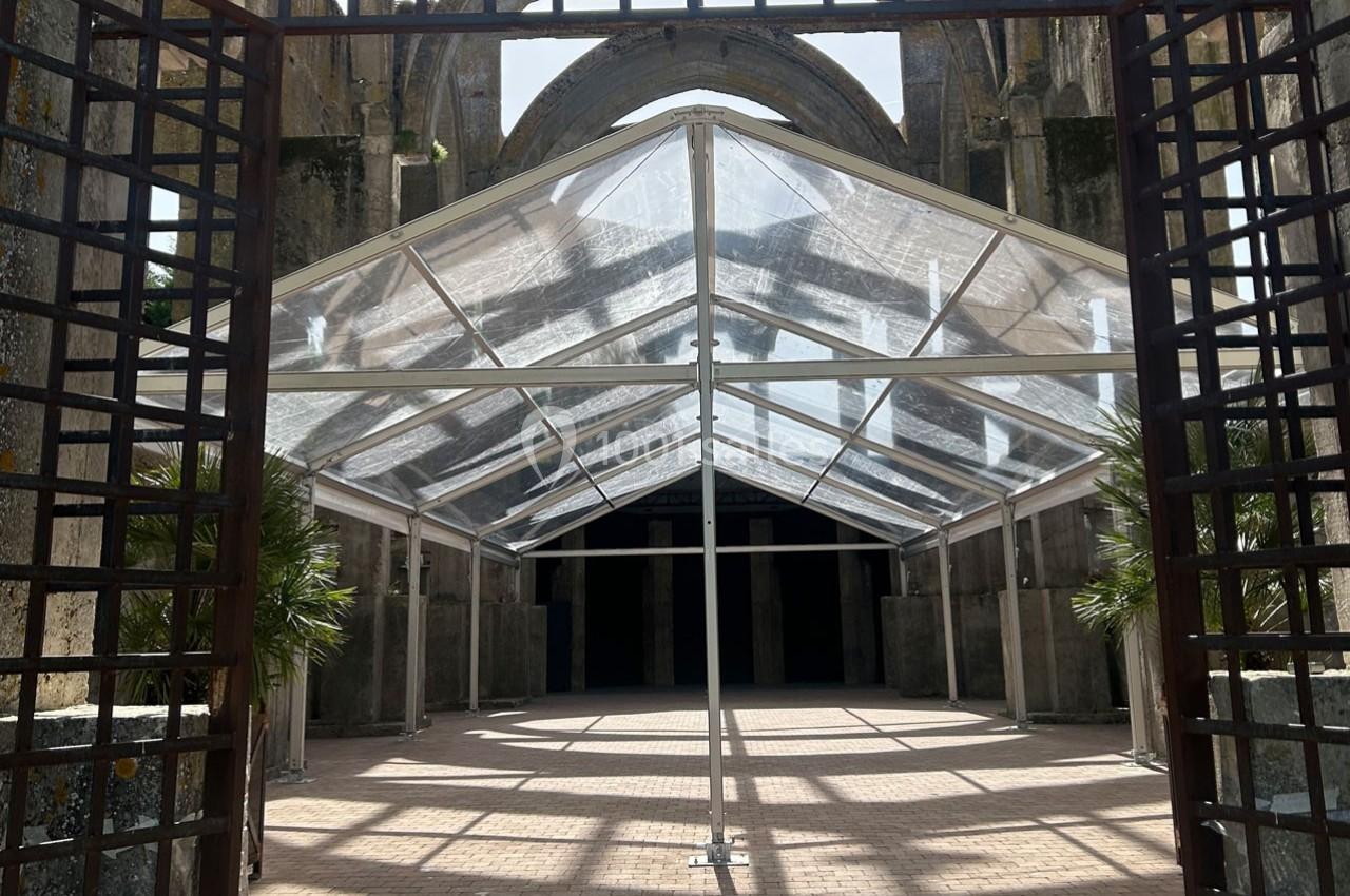 Structure transparente installée sous une ancienne arche en pierre, entourée de grilles métalliques et de végétation.
