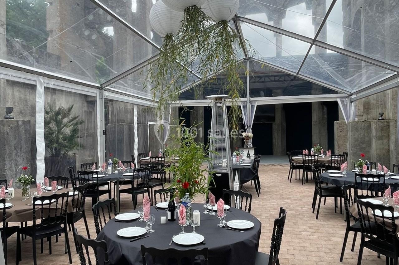 Salle de réception sous une tente transparente, tables rondes dressées avec nappes noires, décorations florales et lanternes…
