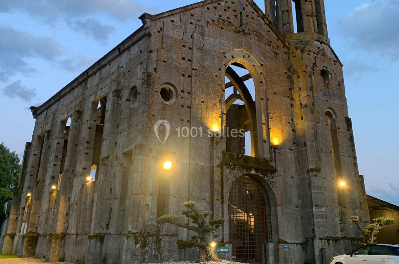 Ruines d'une église en pierre éclairée par des lampes au crépuscule, avec un ciel partiellement nuageux en arrière-plan.