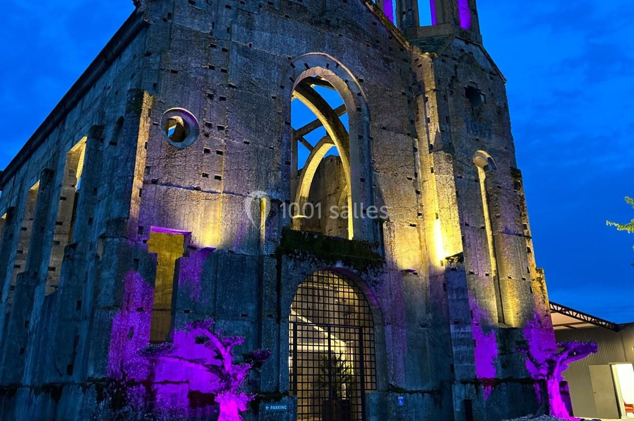 Ruines d'une église illuminées en violet et jaune, avec des sculptures d'arbres à l'avant, sous un ciel crépusculaire.