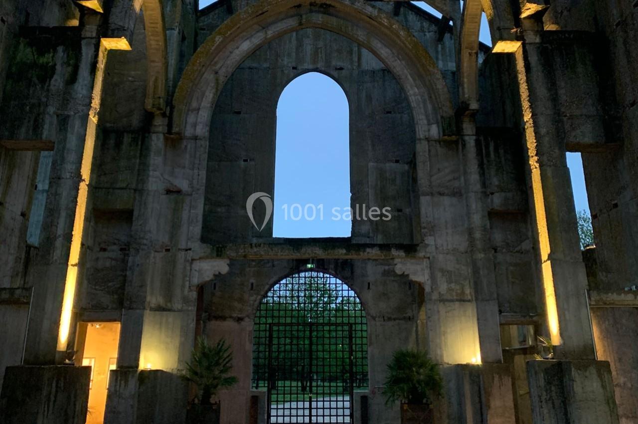Ruines d'un bâtiment avec une grande arche centrale, éclairées par des lumières, donnant sur un portail en fer et un espace…