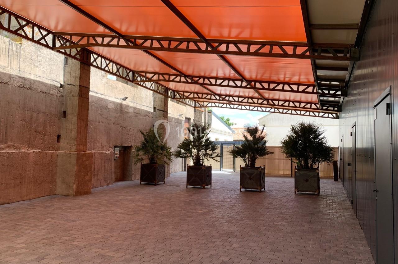 Espace extérieur couvert avec un toit orange, sol pavé et alignement de plantes en bacs devant un mur en béton.